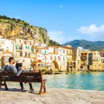 Couple sitting on bench, enjoying view of beach town of Cefalu in Sicily, Italy