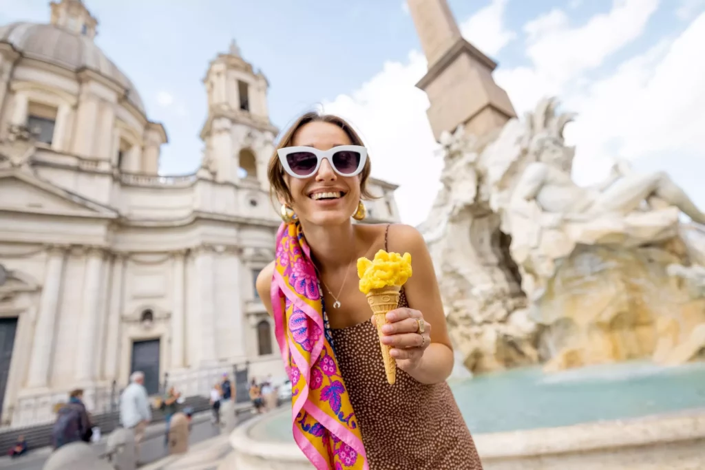Portrait of a cheerful woman eating ice cream in cone while visiting famous Navona square near fountain in Rome. Concept of happy summer vacations, traveling famous italian landmarks