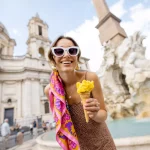 Portrait of a cheerful woman eating ice cream in cone while visiting famous Navona square near fountain in Rome. Concept of happy summer vacations, traveling famous italian landmarks