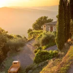 Tuscan countryside stone house with orange three-wheeled pick-up scooter, tall cypress trees, and sunset light flooding distant rolling hills