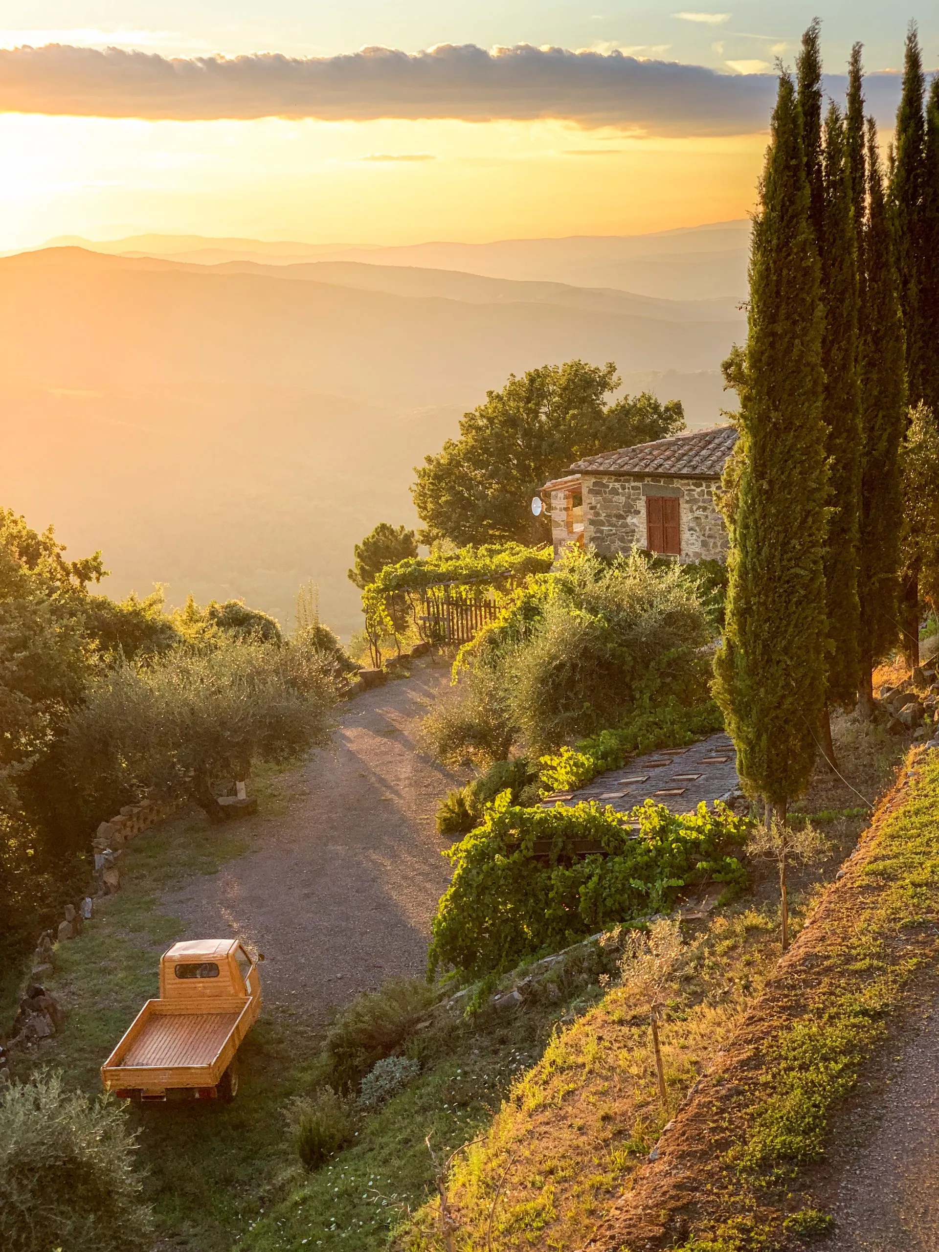 Tuscan countryside stone house with orange three-wheeled pick-up scooter, tall cypress trees, and sunset light flooding distant rolling hills